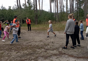 Dzieci z klasy 2b i opiekunowie grają w piłkę na leśnej polanie. Wszyscy uczestnicy są uśmiechnięci, a część dzieci nosi kaski rowerowe.