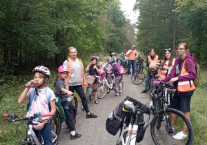 Klasa 2b z wychowawczynią Moniką Mettler i rodzicami podczas rowerowej przerwy w lesie. Dzieci odpoczywają z rowerami, piją napoje, w tle widać oznaczenie szlaku.