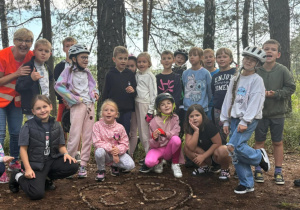 Klasa 2b z wychowawczynią Moniką Mettler podczas wycieczki rowerowej w lesie, przed napisem „2B” ułożonym z szyszek. Dzieci pozują do wspólnego zdjęcia.