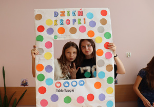 Dwie uczennice klasy 6a ze Szkoły Podstawowej nr 13 w Pabianicach pozują w ramce ozdobionej kolorowymi kropkami z napisem „Dzień Kropki”. Jedna z nich pokazuje znak dłoni, druga uśmiecha się.