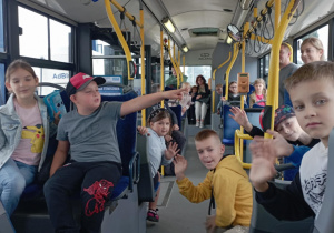 Dzieci z klasy 2b SP13 siedzą w autobusie MZK Pabianice, uśmiechają się i machają do aparatu. W tle widoczni opiekunowie oraz kierowca. Atmosfera radosna i pełna entuzjazmu.