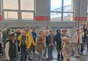 Grupa uczniów klasy 2b SP13 uważnie słucha przewodnika w hali MZK. W tle nauczycielka Monika Juran-Tłoczek, widoczne duże okna i elementy warsztatu.