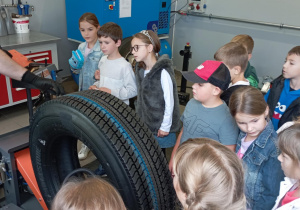 Uczniowie SP13 z klasy 2b podczas wizyty w warsztacie MZK obserwują pokaz pracy wulkanizatora – przyglądają się maszynie i oponie, słuchając wyjaśnień specjalisty.