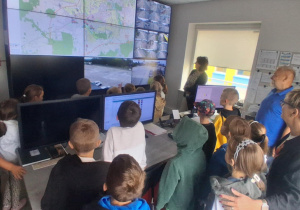 Grupa uczniów klasy 2b obserwuje pracę dyspozytorni MZK, na ekranach widać mapy i monitoring autobusów; z tyłu stoją nauczycielki Monika Juran-Tłoczek i Monika Mettler. Chcesz, żebym teraz przygotował komplet opisów i tekstów alternatywnych dla wszystkich zdjęć w jednym pliku .docx do publikacji lub archiwizacji?