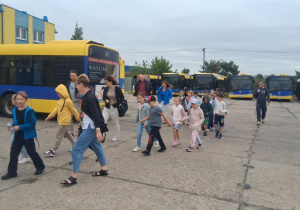 Dzieci z klasy 2b SP13 idą przez zajezdnię MZK wśród żółto-niebieskich autobusów, w tle budynek i opiekunowie.