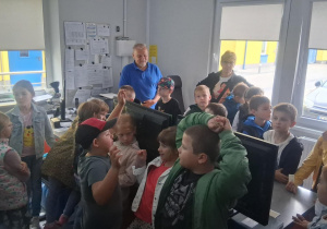 Grupa dzieci z klasy 2b w pomieszczeniu dyspozytorni MZK, słuchają pracownika, wokół stanowiska komputerowe i dokumentacja na ścianach.