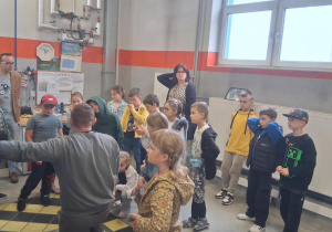 Grupa dzieci z klasy 2b w hali technicznej MZK słucha pracownika prezentującego elementy autobusu; w tle nauczyciele i stanowisko z dokumentami.