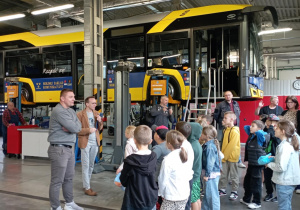 Uczniowie SP13 z klasy 2b zwiedzają warsztat MZK w Pabianicach. W tle podniesione autobusy Solaris. Pracownicy opowiadają dzieciom o tajnikach pracy mechanika i kierowcy.
