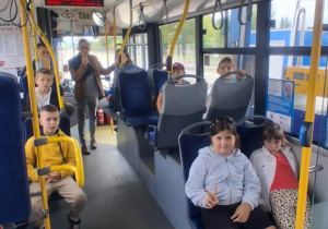 Uczniowie klasy 2B SP13 siedzą w miejskim autobusie Solaris podczas wycieczki do MZK. Uśmiechnięci, w towarzystwie opiekuna, pokazują znak pokoju do aparatu.