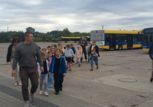 Uczniowie klasy 2B SP13 z nauczycielkami idą w parach po terenie MZK w Pabianicach. W tle widoczne są autobusy w barwach żółto‑niebieskich. Trwa edukacyjna wycieczka.