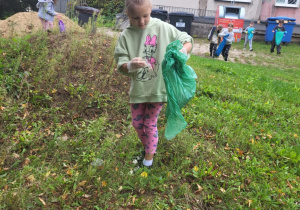 Dziewczynka w zielonej bluzie zbiera śmieci do zielonego worka na terenie zielonym przed szkołą w ramach akcji „Sprzątanie Świata”. W tle widoczne są inne dzieci i kontenery.