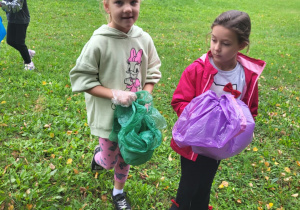 Uczniowie sprzątają teren wokół szkoły podczas akcji „Sprzątanie Świata” – dzieci z kolorowymi workami zbierają śmieci w zarośniętym miejscu przy ogrodzeniu.