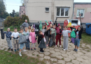 Grupa dzieci z kolorowymi workami na śmieci stojąca przed szkołą po zakończonej akcji sprzątania, w tle kontenery i pojemnik na odzież.