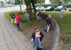 Grupa uczniów sprzątająca teren zielony przy ulicy – dzieci w rękawiczkach trzymają worki na śmieci i zbierają odpady z trawnika i krzewów.