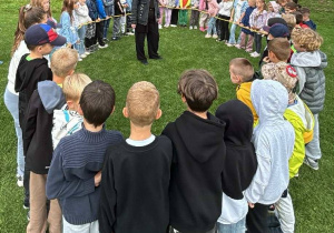 Dzieci stojące w kręgu na trawie podczas zajęć integracyjnych, a w środku nauczycielka prowadząca zabawę na świeżym powietrzu.