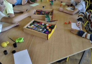 Dzieci siedzące przy stole w świetlicy malują i rysują. Na środku stołu leżą farby, kredki i przybory plastyczne.