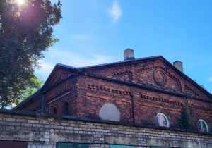 Zabytkowy budynek z czerwonej cegły o przemysłowym charakterze, częściowo zasłonięty przez garaże. W tle błękitne niebo i słońce przebijające przez liście drzewa.