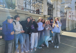 Uczniowie 8a z nauczycielką języka niemieckiego pozują przed zabytkowym ogrodzeniem w słoneczny dzień, w tle brama z kutego żelaza i biały budynek