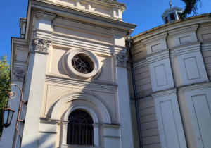 Wieża kościoła ewangelicko-augsburskiego w Pabianicach z zegarem i jasną fasadą na tle błękitnego nieba, cień drzewa pada na budynek.