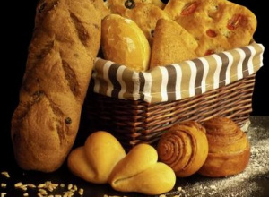 Kosz z różnorodnym pieczywem: chleb, focaccia, bułki i rogaliki, ustawiony na ciemnym tle, otoczony ziarnami i mąką.