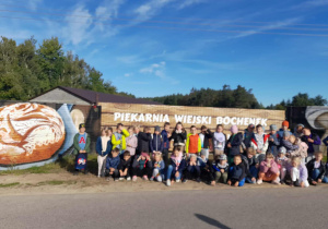 Grupa uczniów przed ogrodzeniem z napisem „Piekarnia Wiejski Bochenek”, obok namalowane bochenki chleba, w tle las i niebieskie niebo.