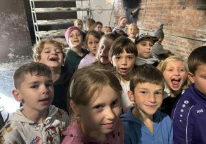 Uczniowie klasy 2A i 2B w piekarni, uśmiechnięci stoją w grupie na tle ceglanej ściany i pieca chlebowego.