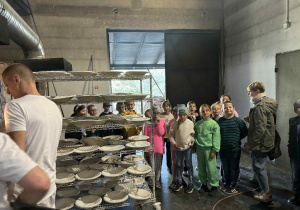 Uczniowie klasy 2A i 2B wraz z nauczycielką Moniką Mettler oglądają półki z uformowanym chlebem w piekarni, czekającym na wypiek.