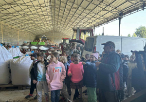 Uczniowie klasy 2A i 2B słuchają rolnika w hali z maszynami i workami zboża podczas wycieczki do gospodarstwa.