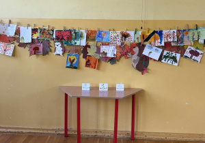 Kolorowe prace plastyczne o tematyce jesiennej zawieszone na sznurku na tle żółtej ściany w świetlicy szkolnej.