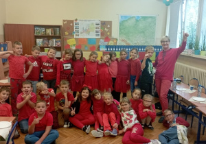 Grupa dzieci z klasy 2b w czerwonych ubraniach pozuje z wychowawczynią Moniką Mettler w klasie podczas szkolnej akcji „Dzień Koloru”. W tle mapy i dekoracje ścienne.
