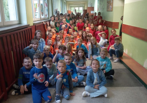 Grupowe zdjęcie uczniów klas I–III SP nr 13 ubranych w kolorowe stroje w ramach szkolnego „Dnia Koloru”, siedzących na szkolnym korytarzu.