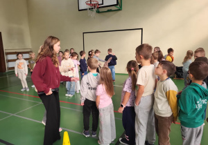 Dzieci z klas I–III w kolejce do zabawy podczas Dnia Integracji w sali gimnastycznej. Starsza uczennica prowadzi zajęcia z mikrofonem.