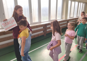 Dziewczynka z tabliczką „Stacja Przytulasy” przytula młodszego chłopca, obok nich stoi uśmiechnięta grupa dzieci – scena z Dnia Integracji w sali gimnastycznej szkoły.