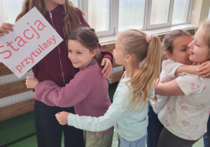 Dziewczynka z tabliczką „Stacja Przytulasy” przytula młodszego chłopca, obok nich stoi uśmiechnięta grupa dzieci – scena z Dnia Integracji w sali gimnastycznej szkoły.