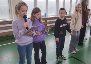 Dzieci stojące w szeregu na sali gimnastycznej – dziewczynka mówi do mikrofonu, inne dzieci trzymają kartki i słuchają.