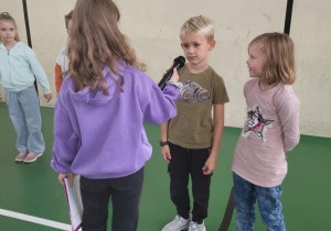 Dziewczynka trzyma mikrofon przed chłopcem, obok stoi dziewczynka – dzieci biorą udział w szkolnym wydarzeniu integracyjnym na sali gimnastycznej.