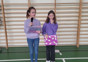 Dwie uczennice prowadzące wydarzenie szkolne stojące przy drabinkach gimnastycznych; jedna z nich mówi do mikrofonu.