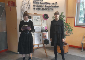 Dwie uczennice w czarnych strojach stoją w holu szkoły przy gazetce poświęconej patronce Stefanii Sempołowskiej. Jedna trzyma kosz z cukierkami, obok widoczne są prace plastyczne.