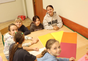 Uczniowie klasy 4 siedzą przy stole i piszą list do patronki szkoły. Pracują nad kolorowym arkuszem papieru, uśmiechając się w stronę aparatu w sali lekcyjnej.