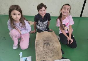 Troje dzieci siedzi na sali gimnastycznej obok dużego arkusza papieru z rysunkiem postaci i napisem „KUKAIGAHLA”. Uśmiechnięte pozują do zdjęcia po zakończonej pracy plastycznej.