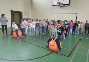 Uczniowie klas I–III podczas zawodów w sali gimnastycznej. Dwoje dzieci skacze na dużych pomarańczowych piłkach z buzią kota, a reszta uczniów stoi w rzędach i kibicuje.