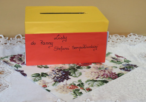 Kolorowe pudełko z napisem „Listy do Panny Stefanii Sempołowskiej” ustawione na ozdobnym obrusie z koronką i kwiecistym wzorem, służące do zbierania listów od uczniów.