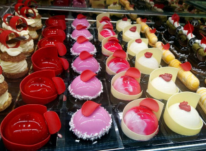 Witryna cukierni z rzędem kolorowych deserów — różowych, czerwonych i białych musów w błyszczących polewach, ozdobionych dekoracjami z czekolady i owoców, ustawionych w eleganckich formach.