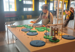 Uczeń przy stole z zielonymi i niebieskimi kołami zębatymi wykonuje eksperyment w Centrum Nauki EC1 w Łodzi.