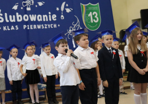 Chłopiec w białej koszuli trzyma mikrofon i recytuje wiersz, obok stoją inni uczniowie w eleganckich strojach.