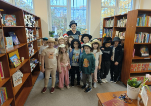 Grupa uczniów klasy 1 stoi w bibliotece szkolnej wraz z wychowawczynią. Dzieci mają na głowach różnorodne kapelusze — słomkowe, materiałowe i ozdobne — w ramach „Dnia kapelusza”. W tle widać regały pełne książek.