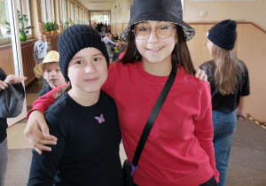 Dwójka uczniów pozuje na szkolnym korytarzu podczas „Dnia kapelusza”. Jedno dziecko ma czarną czapkę zimową, drugie kapelusz typu bucket. W tle widać innych uczniów również w różnych nakryciach głowy.