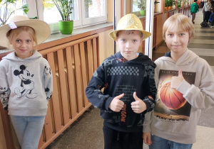 Troje uczniów pozuje na szkolnym korytarzu podczas „Dnia kapelusza”. Dziewczynka po lewej ma szeroki słomkowy kapelusz, chłopiec w środku złoty błyszczący kapelusz, a chłopiec po prawej stoi bez nakrycia głowy. W tle widać innych uczniów oraz szkolne okna.