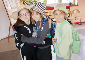 Trzy uczennice klasy 2 stoją na szkolnym korytarzu podczas „Dnia kapelusza”. Przytulają się i pozują do zdjęcia w fantazyjnych kapeluszach — słomkowym, w biało-czarne pasy oraz szydełkowym. W tle widać rośliny i szkolną dekorację.