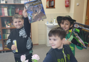 Chłopiec dumnie trzyma w górze dużą książkę „Wielka Encyklopedia Zwierząt”, obok drugi uczeń siedzi przy biurku z zakładką w ręku, a w tle dziecko w kapeluszu trzyma kolejną książkę. Scena odbywa się w bibliotece szkolnej podczas pasowania pierwszoklasistów na czytelników.